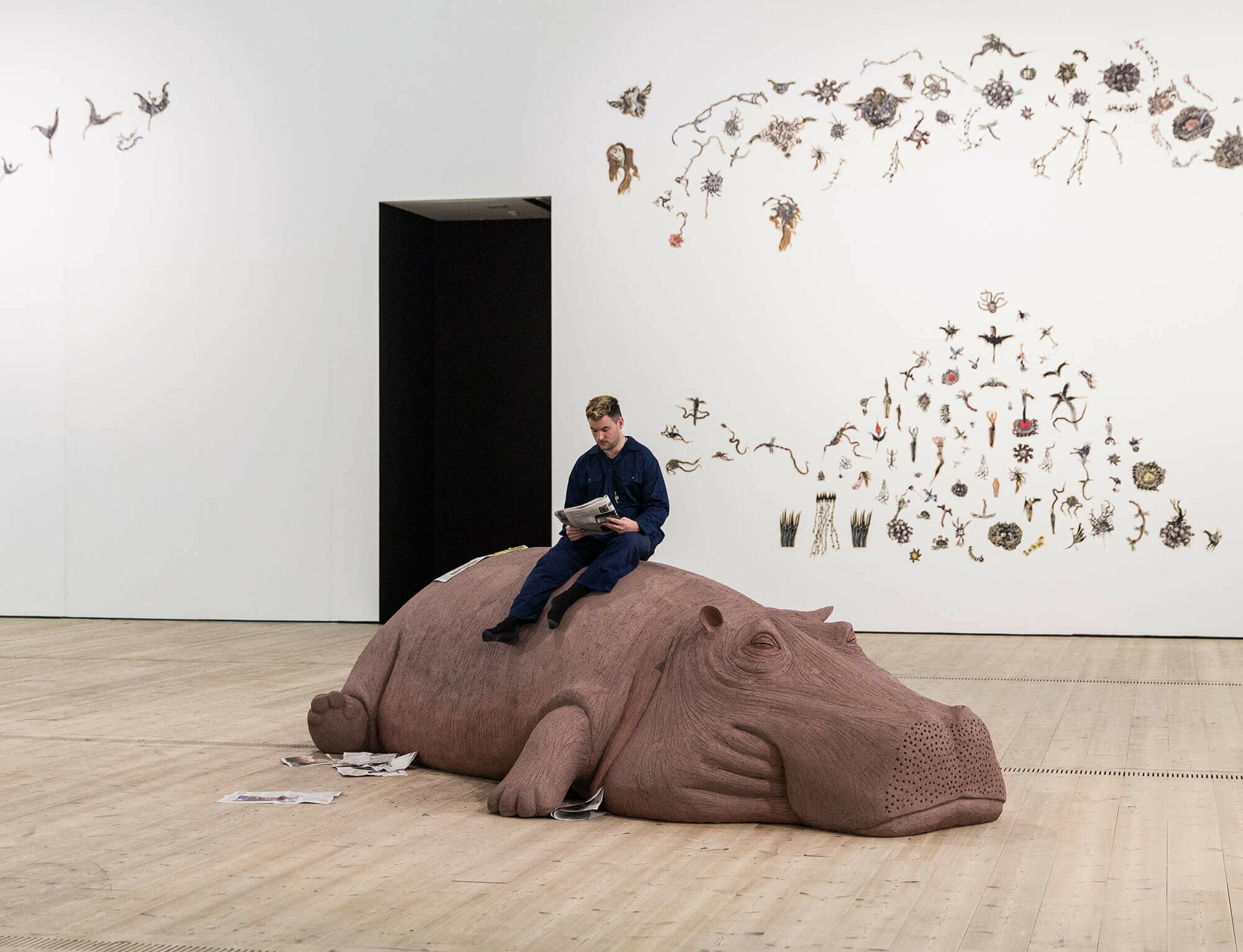 Man sits reading on a sculptural hippo inside a exhibition space.